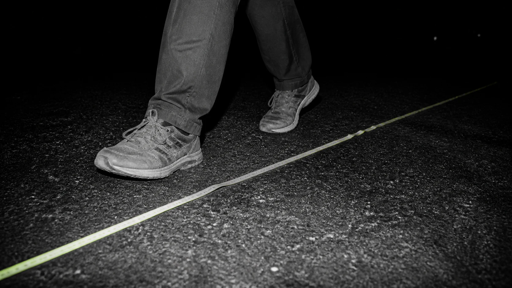 Black-and-white flash photograph of worn walking shoes beside a measuring tape on asphalt