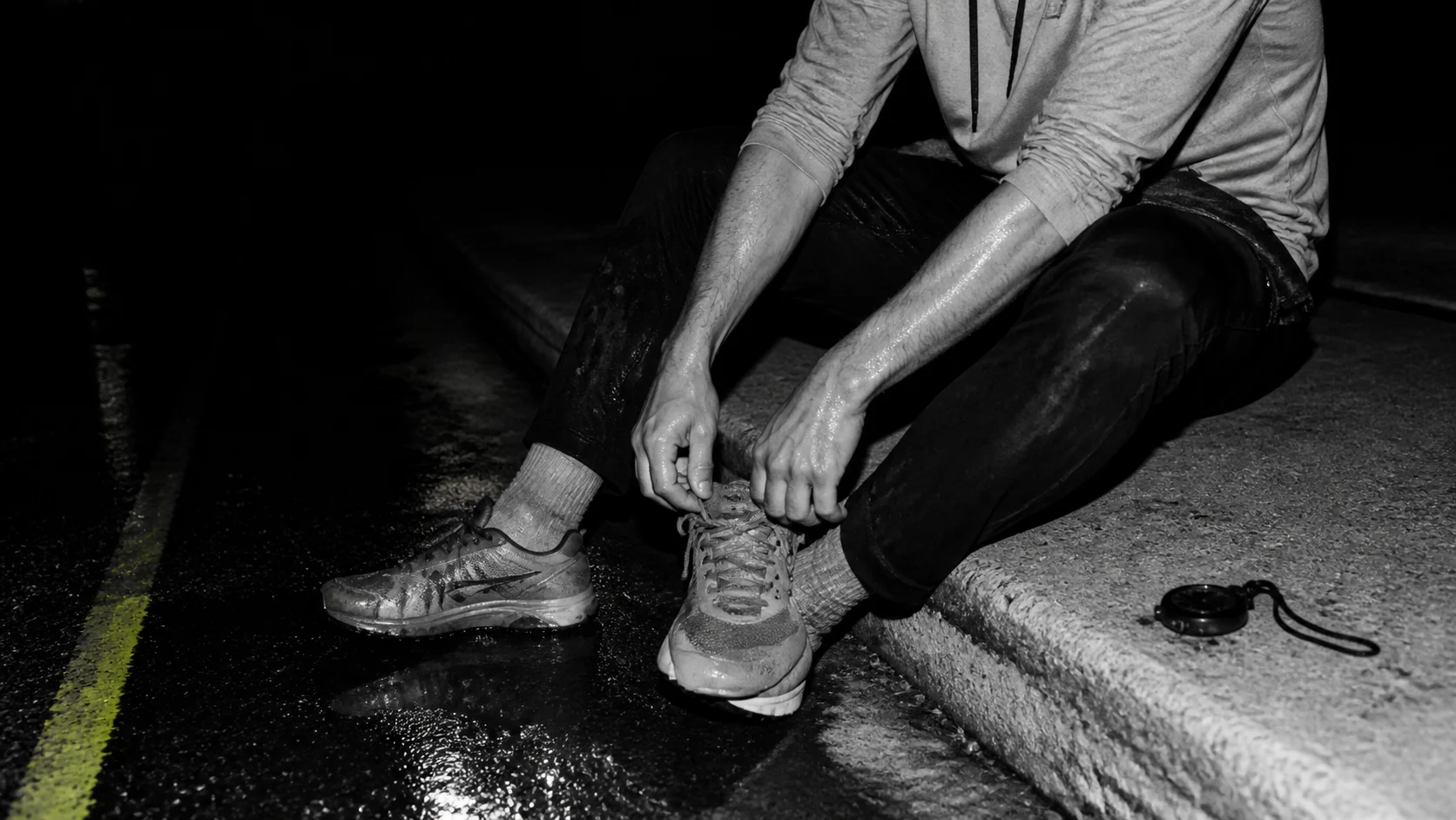 Black-and-white flash photograph of a walker adjusting a wet shoe beside a dark outdoor track